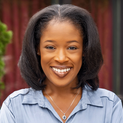 Professional headshot of Sydne Lewis, a smiling woman with straight black hair, wearing a light blue shirt and a silver necklace, set against a blurred outdoor background.